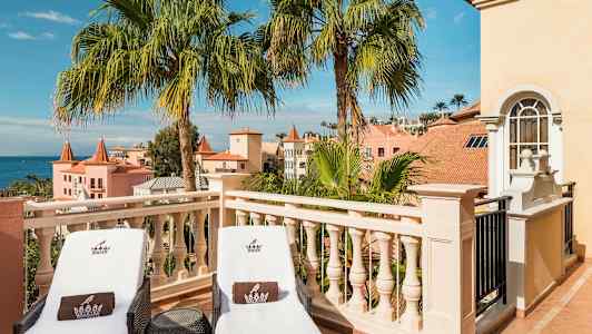 To solsenger på en terrasse med utsikt over palmer og pastellfargede bygninger. (AI-generert tekst)