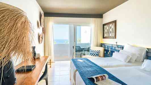 Et lyst hotellrom med to senger, en trebenk og en balkong med havutsikt. (AI-generert tekst)