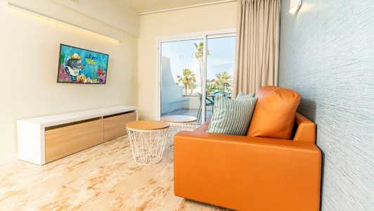 Moderne stue med oransje sofa, puter, sofabord og TV samt utsikt mot balkong og palmeskog. (AI-generert tekst)