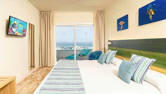 Hotellrom med to senger, stripete sengetepper og blå puter, en balkong med utsikt mot havet. (AI-generert tekst)