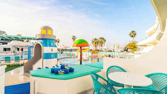 Et basseng med solsenger og et lekeområde med vannleker, inkludert en fargerik fyrformet sklie. (AI-generert tekst)