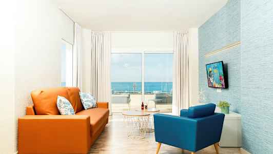 Et oppholdsrom med en oransje sofa og en blå lenestol foran et bord. Et stort vindu i bakgrunnen viser utsikt over havet. (AI-generert tekst)