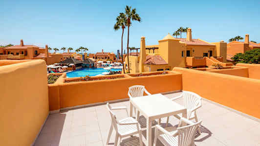 En terrasse med bord og stoler, med utsikt over et basseng og omkringliggende bygninger. (AI-generert tekst)