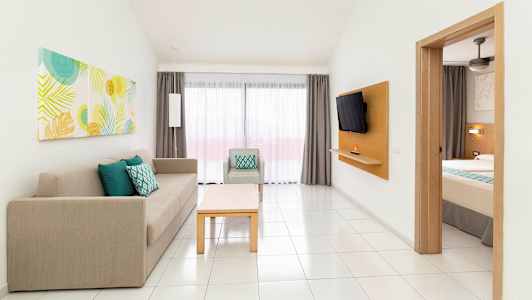 Et hotellrom med en delvis synlig seng, en sofa og lenestol samt et bord. Et vindu med gardiner er synlig i bakgrunnen. (AI-generert tekst)