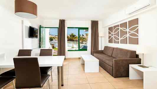 Et lyst og moderne innredet hotellrom med en brun sofa, et spisebord og stoler. Vinduer og dør leder til utendørsområdet. (AI-generert tekst)