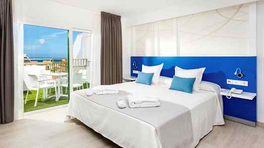 Bilde av et hotellrom med en dobbeltseng, blå ryggstøtte og et vindu som viser en balkong med møbler og havutsikt. (AI-generert tekst)