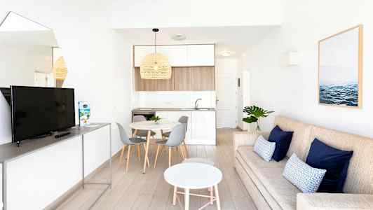 En moderne stue med en beige sofa, blå puter, et hvitt rundt salongbord og en TV på stativ. Ved siden av er det et spiseområde med et rundt bord og grå stoler samt et kjøkken i lyse farger. (AI-generert tekst)