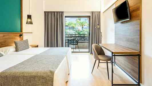 Hotellrom med to senger, skrivebord, TV og balkong med utsikt over trær og grøntområder. (AI-generert tekst)