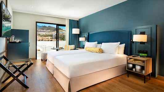 Et hotellrom med to enkeltsenger, blå vegger og en balkong med utsikt mot fjellene. (AI-generert tekst)