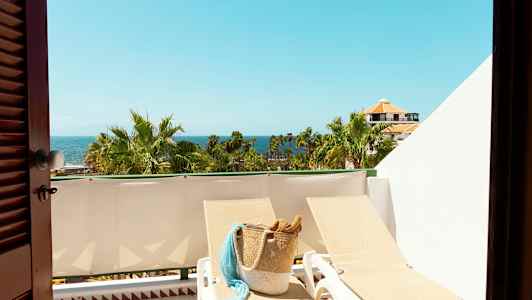 Utsikt over en solrik balkong med solsenger, en kurvveske, palmer og havet i bakgrunnen. (AI-generert tekst)