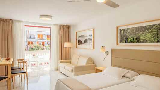 Et lyst hotellrom med to senger, en sofa og spisebord med stoler, samt et stort vindu med utsikt over balkongen og bygninger utenfor. (AI-generert tekst)