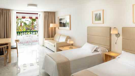 Hotellrom med to enkeltsenger, en sofa, et bord med stoler og et vindu mot en balkong. (AI-generert tekst)