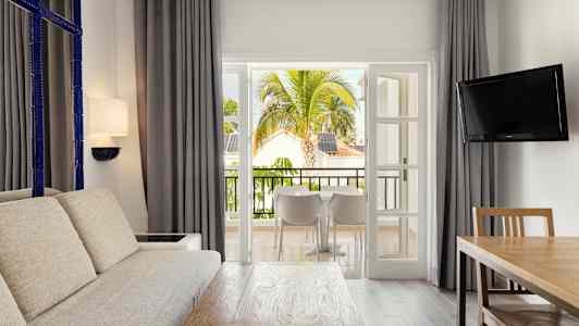 Et oppholdsrom med stor sofa, trebord og en TV, åpner opp mot en balkong med utsikt over tropiske trær og bygninger. (AI-generert tekst)