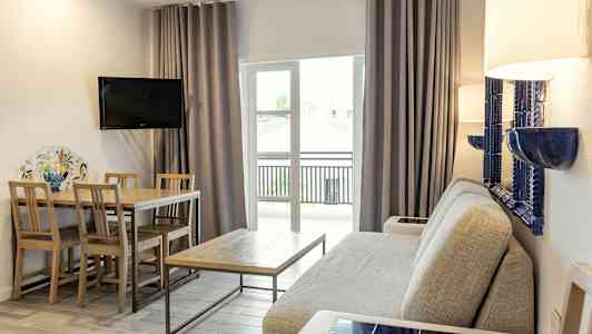 En stue med en grå sofa, et trebord med stoler, en veggmontert TV og glassdører med gardiner. (AI-generert tekst)