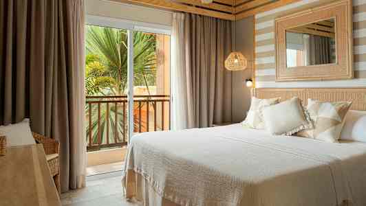 Et hotellrom med en dobbeltseng, lyse tekstiler og interiørdetaljer. Utenfor er det en balkong med utsikt over grøntområder. (AI-generert tekst)