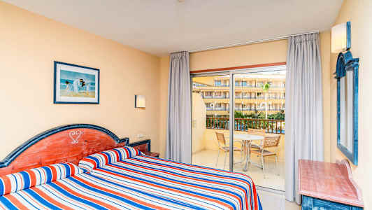 Hotellrom med to enkeltsenger, stripete blå, røde og hvite sengetepper, og utsikt mot en balkong med møbler. (AI-generert tekst)