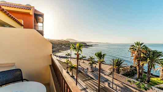 Utsikt fra en balkong mot en palmekantet strandpromenade og havet under en klar himmel. (AI-generert tekst)