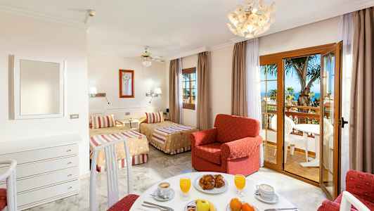 Et hotellrom med to senger, en rød lenestol og en spiseplass, samt en balkong med utsikt over palmer og havet. (AI-generert tekst)