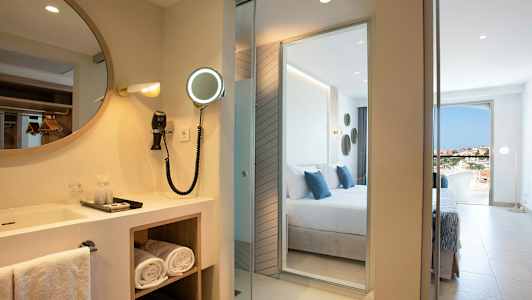 Hotellrom med et speil og vask ved siden av et soverom med utsikt. (AI-generert tekst)