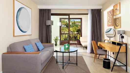 Et hotellrom med en grå sofa, et skrivebord og en balkong med utsikt over en hage. (AI-generert tekst)