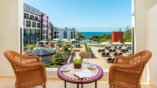 Utsikt fra en balkong over et hotellbasseng med solsenger og havet i bakgrunnen. (AI-generert tekst)