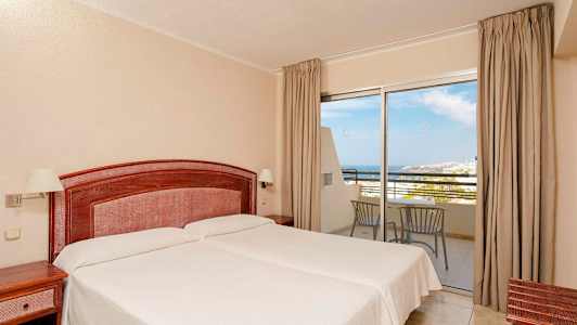 Et hotellrom med to enkeltsenger, en trehodegavl og nattbord. Et glassparti med utsikt mot havet og balkongmøbler. (AI-generert tekst)