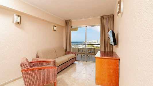 En stue med en sofa, en lenestol, et trebord og en veggmontert TV. I bakgrunnen er det en balkong med stoler og utsikt mot havet. (AI-generert tekst)