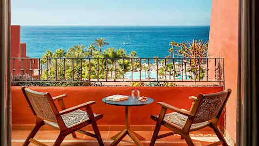 To stoler og et bord på en terrasse med utsikt over et basseng, palmer og havet. (AI-generert tekst)