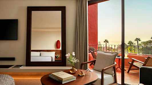 Et elegant hotellrom med et speil, åpning mot en møblert terrasse og havutsikt. (AI-generert tekst)