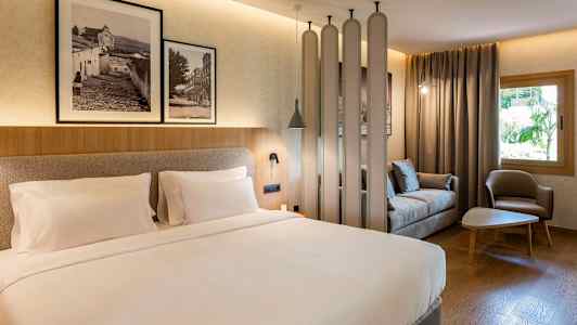 Et hotellrom med en dobbeltseng, en sittegruppe med sofa og lenestol, samt dekorative detaljer som bilder på veggen. (AI-generert tekst)