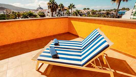 Utsikt fra en solterrasse med to solsenger med stripete blå og hvite puter samt håndklær på dem, omgitt av palmer og bygninger. (AI-generert tekst)