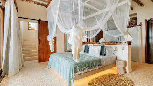 Et soverom med himmelseng med hvitt myggnett, lyse vegger og tregulv. En trapp er synlig i bakgrunnen. (AI-generert tekst)