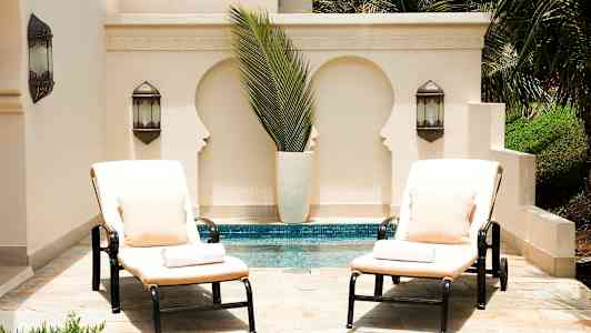 To solsenger med puter og puter står ved et basseng foran en lys vegg med dekorativt utskjæringer og grønne planter. (AI-generert tekst)