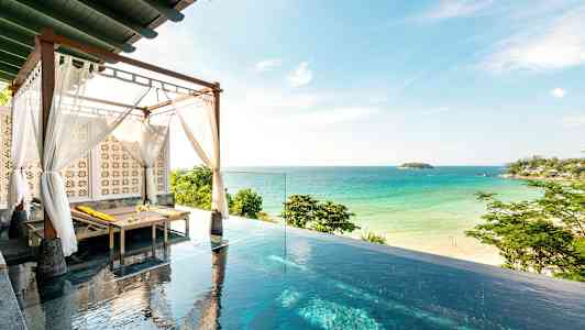 Bildet viser et infinitybasseng med en solseng under et tak med hvite tekstiler, og i bakgrunnen er en sandstrand og havet. (AI-generert tekst)