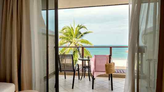 Utsikt fra hotellrom til en balkong med to stoler og et bord, samt en strand og havet i bakgrunnen. (AI-generert tekst)