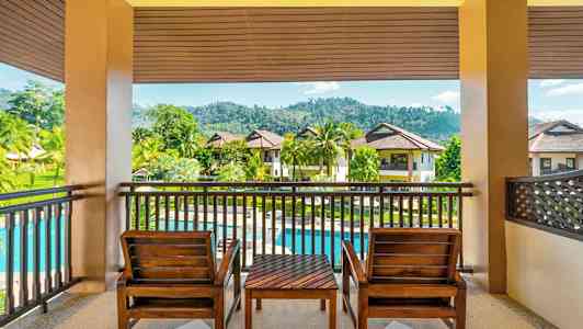 En terrasse med to trestoler og et bord med utsikt over en grønn hage, et basseng og flerbolighus. (AI-generert tekst)