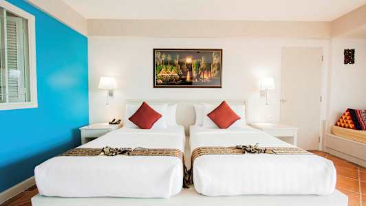 Et hotellrom med to enkeltsenger, hvitt sengetøy og dekorative røde puter. På veggen henger et fargerikt maleri. (AI-generert tekst)