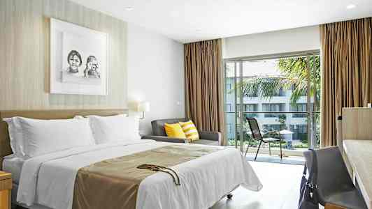 Et hotellrom med seng, sofa med fargerike puter og bord med stol ved et vindu som vender mot en balkong med utsikt over en hage. (AI-generert tekst)