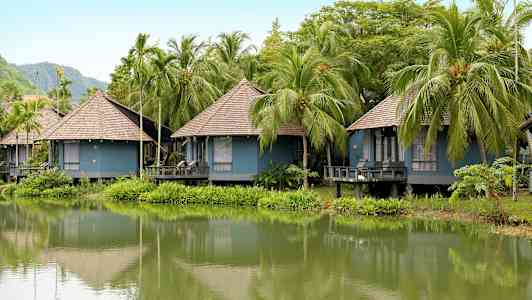 Flere blå bungalower med stråtak ligger ved en frodig innsjø omgitt av palmer. (AI-generert tekst)