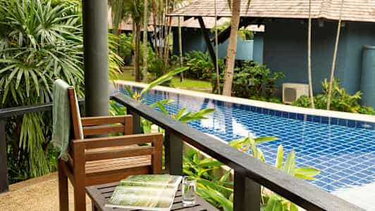 Utsikt fra en veranda mot et basseng omgitt av grønne planter og trær. (AI-generert tekst)