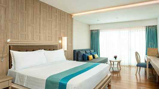 Et hotellrom med en dobbeltseng, en sofa med puter, et bord og en skrivebordsstol. Rommet har trepaneler på veggene og gulv, samt store vinduer med gardiner. (AI-generert tekst)