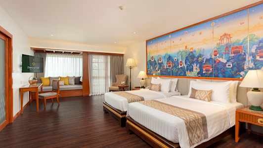 Et hotellrom med to senger, et stort fargerikt kunstverk på veggen og en sitteområde med vindu. (AI-generert tekst)