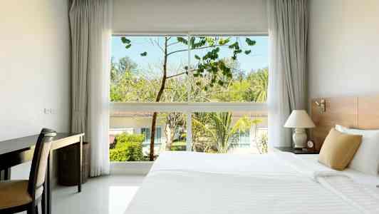 Et hotellrom med en stor seng, et skrivebord og et vindu som viser trær og bygninger utendørs. (AI-generert tekst)