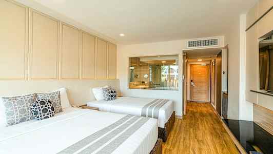 Et hotellrom med to brede senger oppredd med hvitt sengetøy og dekorative puter. Gulvet er av tre, og i bakgrunnen er det en glassvegg med utsyn til badet. (AI-generert tekst)