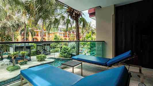 Utsikt fra en balkong med to blå solsenger som vender mot en fargerik hage med palmer og basseng. (AI-generert tekst)