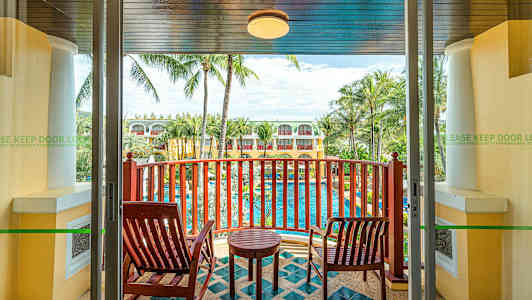 Utsikt fra en balkong med to trestoler, et lite bord og en utsikt over et basseng omgitt av palmer og bygninger. (AI-generert tekst)