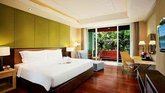 Soverom med en stor seng, sittegruppe og skrivebord, samt utgang til en balkong som har utsikt over en hage. (AI-generert tekst)