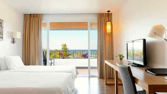 Et hotellrom med to senger, skrivebord og utsikt mot en balkong med hav i bakgrunnen. (AI-generert tekst)