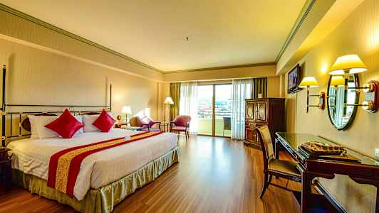 Et hotellrom med en dobbeltseng og rød dekor, sittegruppe ved balkongdøren, og skrivebord med speil. (AI-generert tekst)