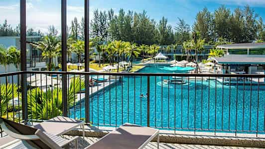 Et basseng omgitt av bygninger og palmer, sett fra en veranda med solsenger. (AI-generert tekst)